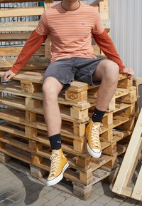 Camiseta de rayas naranja y crema, mangas largas rojas, pantalones cortos grises, zapatillas de caña alta amarillas, calcetines negros, sentado sobre palets de madera.