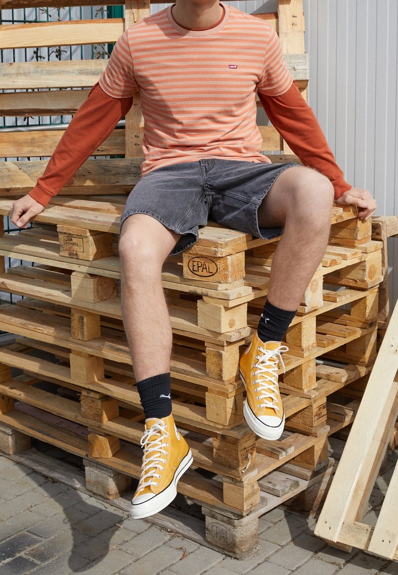 Camiseta de rayas naranja y crema, mangas largas rojas, pantalones cortos grises, zapatillas de caña alta amarillas, calcetines negros, sentado sobre palets de madera.