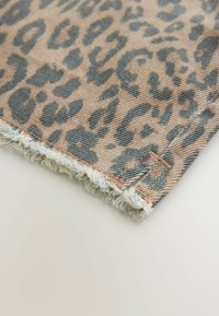 Leopardmönstrad denimtyg med en tanbakgrund, som har en fransad fåll och mörkblå svarta fläckar. Den strukturerade ytan visar tygväven.