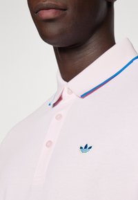 Polo shirt rosa claro com colarinho canelado e detalhes em azul. Três botões na parte da frente e um pequeno logótipo azul bordado.