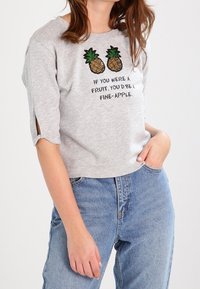 Camiseta corta gris de algodón, con piñas bordadas y texto. Mangas cortas con aberturas para un detalle adicional. Combinada con vaqueros azules.
