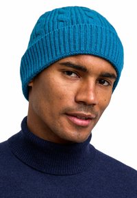 Teal knitted beanie with ribbed texture, featuring a subtle cable pattern. Paired with a dark blue turtleneck sweater.