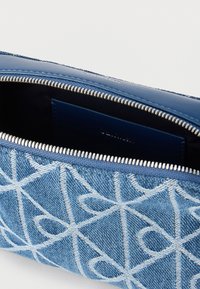 Μπλε τσαντάκι από τζιν με λευκή γεωμετρική ραφή, μερικώς ανοιχτό φερμουάρ που αποκαλύπτει σκοτεινό εσωτερικό και ένα μικρό δερμάτινο θήκη καρτών μέσα.