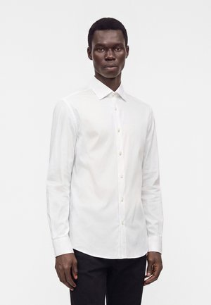 Homme de face portant une chemise blanche à manches longues boutonnée et un pantalon noir sur un fond blanc uni.