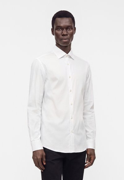 Uomo rivolto in avanti che indossa una camicia bianca a maniche lunghe con bottoni e pantaloni neri su uno sfondo bianco semplice.