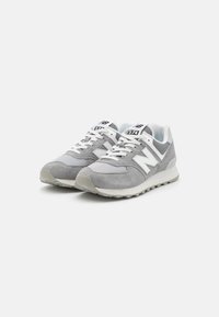 Pár šedých a bílých tenisek New Balance 574 s horní částí z prodyšné síťoviny a semiše, bílými tkaničkami a texturovanou gumovou podrážkou na bílém pozadí.