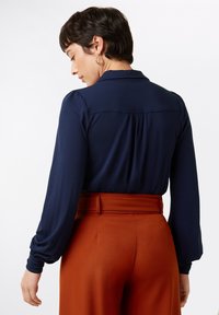 Marinblå långärmad blus med draperad ryggdetalj, kombinerad med rostfärgade höga byxor. Slät tyg med avslappnad passform.