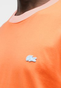 Κοντομάνικη πορτοκαλί μπλούζα με αντίθετο ανοικτό ροζ γιακά και ανοιχτό μπλε κεντημένο λογότυπο στο στήθος. Ομαλή υφή βαμβακιού.