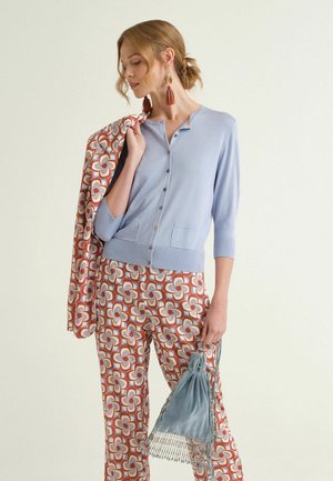 Donna che indossa un cardigan azzurro chiaro con bottoni e pantaloni con motivi, tiene una giacca abbinata sulla spalla e una borsa con frange blu e rosa.