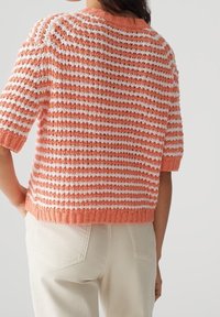 Mujer vestida con un suéter de punto texturizado de manga corta, con rayas horizontales en color coral y blanco, combinado con pantalones de color beige claro.