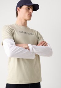 Béžové tričko s logem "calvin klein", vrstvené přes bílé tričko s dlouhým rukávem. Model má na sobě námořnickou baseballovou čepici. Bavlněná směs, ležérní design.