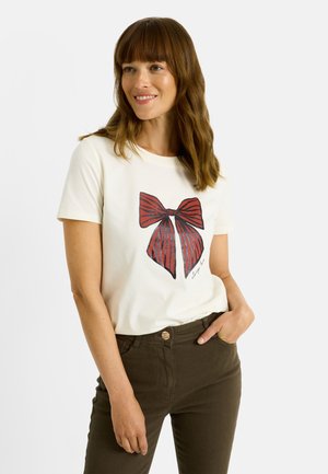 Camiseta blanca de manga corta con un gran gráfico de un lazo rojo y rayas azules, combinada con pantalones verde oscuro. Tejido suave, ajuste casual.