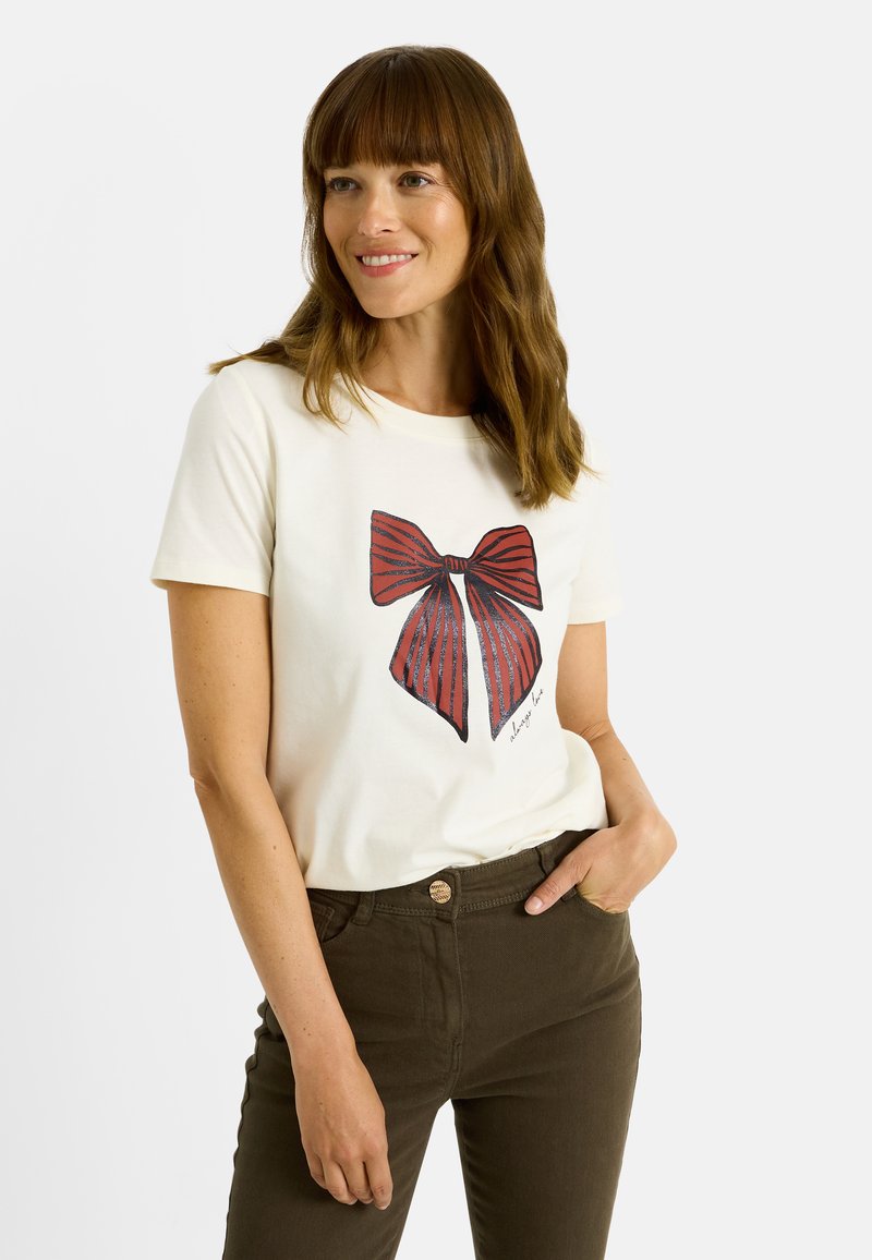 Camiseta blanca de manga corta con un gran gráfico de un lazo rojo y rayas azules, combinada con pantalones verde oscuro. Tejido suave, ajuste casual.