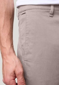 Beige katoenen broek met een gladde textuur, voorzien van zijzakken en een rechte snit. Hand die de tailleband vasthoudt toont de details van de stof.