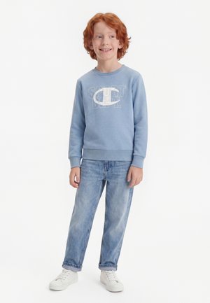 Lächelnder rothaariger Junge, der einen blauen Pullover mit dem Text "SPORT DEPT.", hellblaue Jeans und weiße Turnschuhe trägt und vor einem weißen Hintergrund steht.