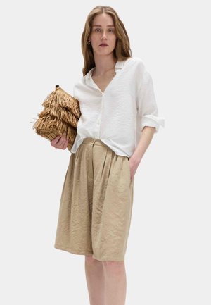 Donna che indossa una camicia bianca con bottoni e maniche arrotolate e pantaloncini beige a pieghe fino al ginocchio, che tiene in mano una pochette marrone con texture.