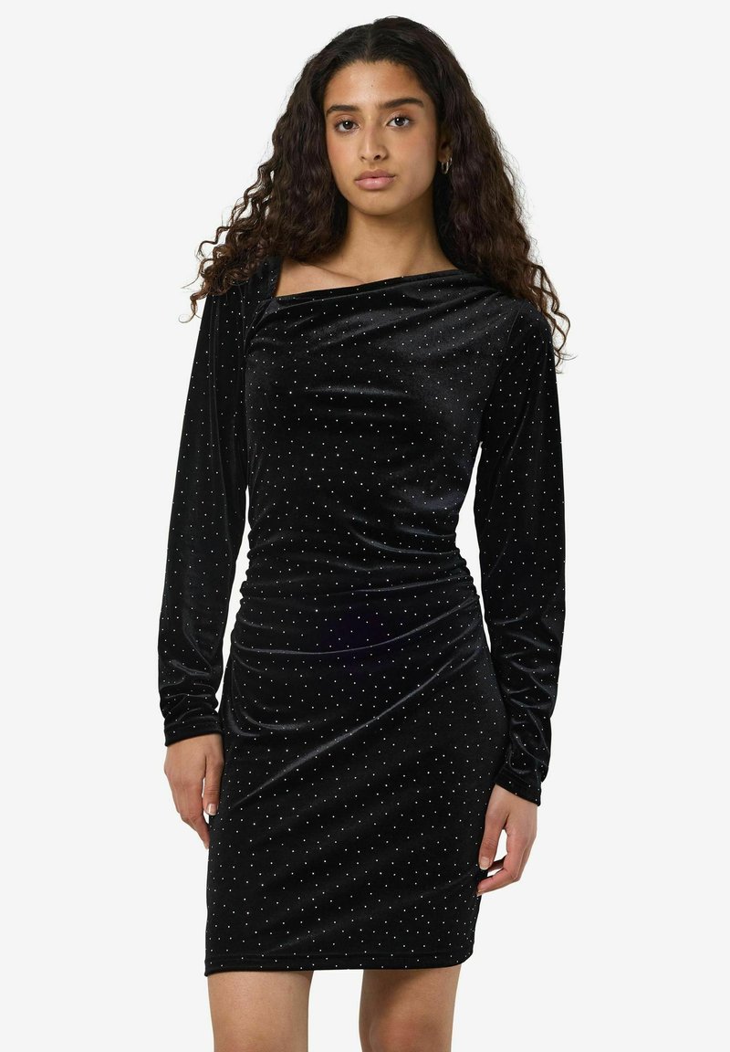 Svart fløyelskjole med en tettsittende silhuett, lange ermer og en båthals. Den har spredte sølvfargede prikker og rynkedetaljer ved midjen.