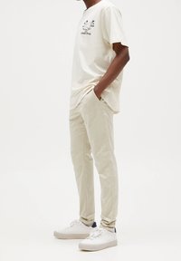 Uomo in piedi con le mani nelle tasche, che indossa pantaloni beige, una maglietta grafica chiara e sneakers bianche su uno sfondo bianco.