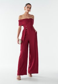 Červený overal so zložkou odhalených ramien, textúrovanou látkou, širokými nohavicami, pásom s pozlátenými ozdobami a elegantnými rukávmi.