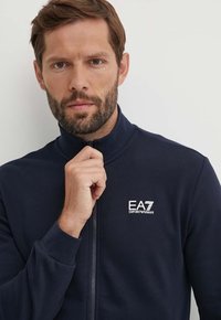 Giacca zip-up navy realizzata in tessuto morbido. Presenta un colletto alto e il logo EA7 ricamato sul lato sinistro del petto. Texture liscia e design aerodinamico.