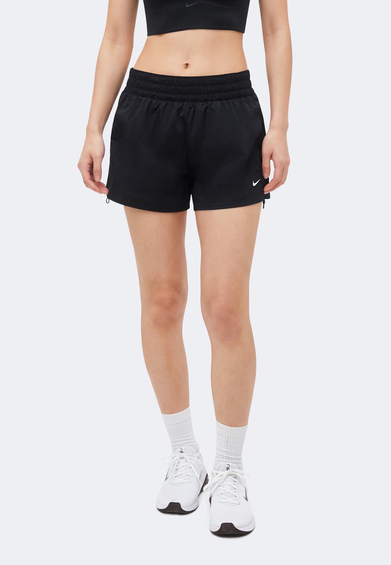 Γυναίκα φοράει μαύρα αθλητικά σορτς Nike, λευκές κάλτσες και λευκά παπούτσια τρεξίματος Nike και στέκεται μπροστά σε απλό φόντο.