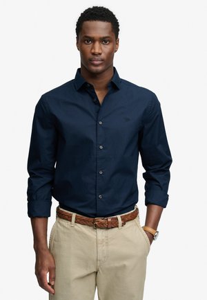 Mężczyzna w granatowej koszuli na guziki z podwiniętymi rękawami, beżowych spodniach, brązowym plecionym pasku i zegarku na lewym nadgarstku stoi z rękami w kieszeniach.