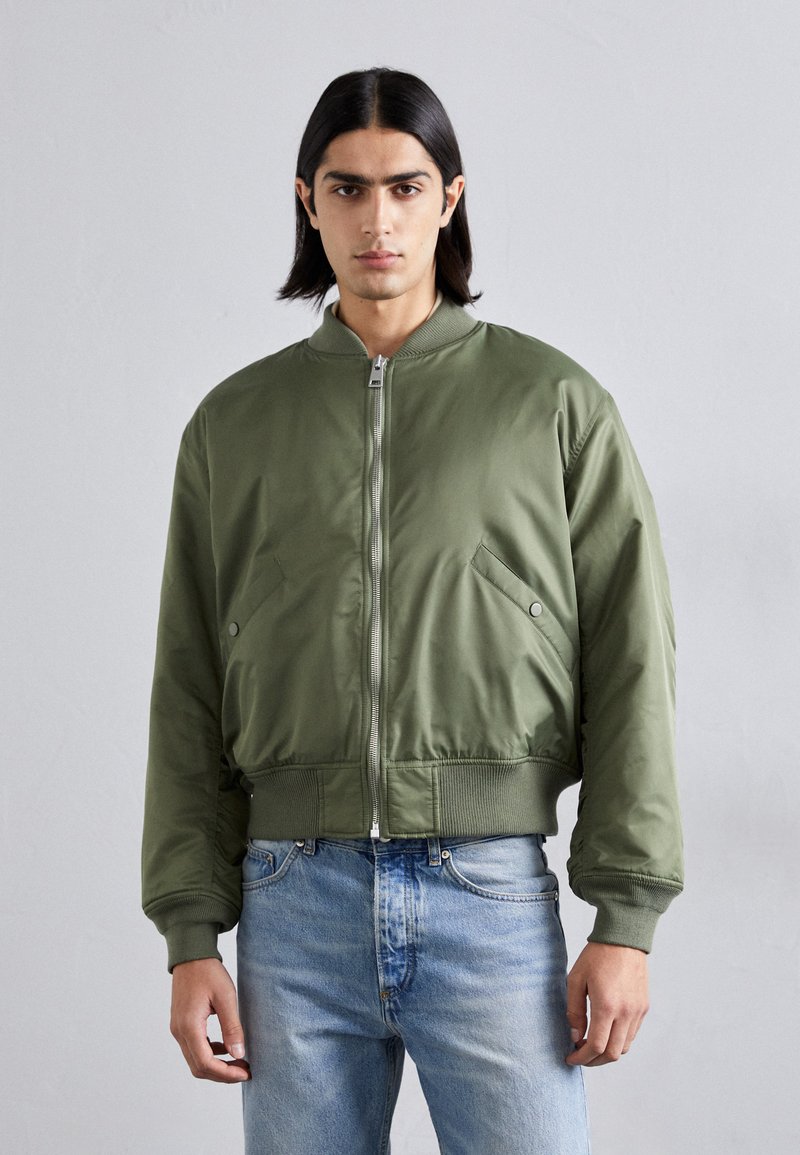 Chaqueta bomber verde oliva con cremallera frontal, puños acanalados y dos bolsillos laterales; combinada con jeans de mezclilla azul claro. Textura suave.