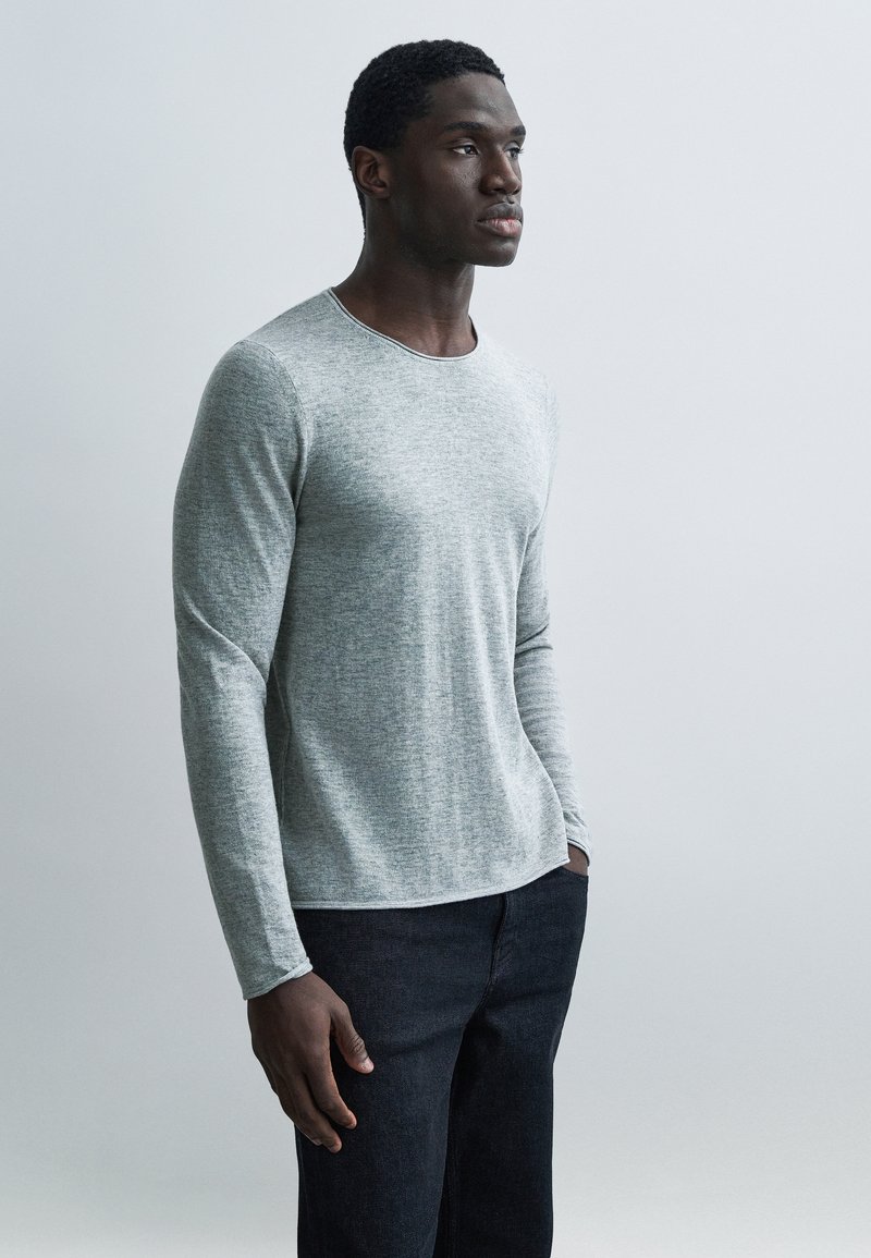 Γκρι μακρυμάνικο πουκάμισο με στρογγυλή λαιμόκο ψη, κατασκευασμένο από υφή υφάσματος, φορεμένο με σκούρο τζιν, τα χέρια στις τσέπες, μπροστά σε φωτεινό φόντο.