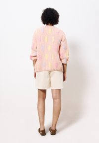 Pletený svetr v pastelově růžové barvě s texturovaným vzorem a barevnými akcenty. Má volný střih, balónové rukávy a žebrovaný lem. Nošený s béžovými šortkami.