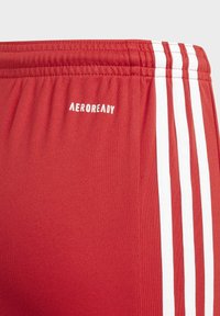 Shorts de sport rouges avec des bandes blanches sur les côtés, fabriqués à partir d'un tissu lisse et extensible. Présentent le logo "AEROREADY" sur la ceinture.