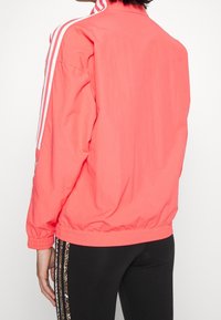 Person klädd i en korallrosa jacka med vita ränder på ärmarna och svarta leggings med mönstrade sidostripes, visad bakifrån.