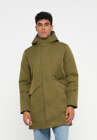 Mann trägt einen olivgrünen Winterpark mit Kapuze und Klapptaschen sowie hellblauen Jeans, steht vor einem weißen Hintergrund.