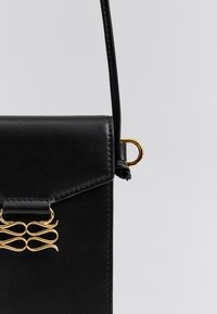 Μαύρη δερμάτινη τσάντα χειρός με δομημένο σχέδιο, έντονα χρυσά μεταλλικά στοιχεία και διακοσμητικό κυματιστό διακοσμητικό κάτω από το κούμπωμα του καπακιού.