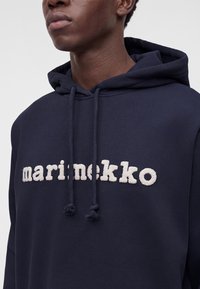 Homme portant un sweat à capuche bleu marine foncé avec un texte blanc texturé "marimekko" et des cordons assortis sur un fond uni.