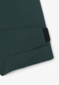 Donkergroene kleding met een opgevouwen manchet, voorzien van een zwarte klittenbandsluiting en een embossed groen krokodillogo op de stof.