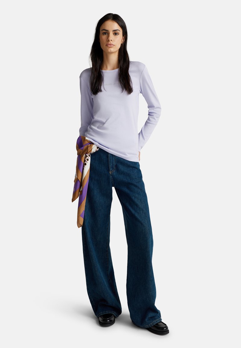 Frau trägt ein helllila langärmliges Oberteil, dunkle weite Jeans, schwarze Schuhe und einen lila-braun gemusterten Schal, der in der Taille gebunden ist.