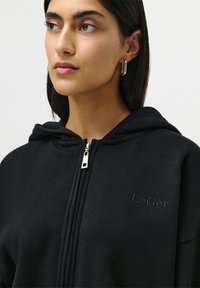 Woman with long dark hair wears silver hoop earrings and a black zip-up hoodie embroidered with "LeGer" on the chest.