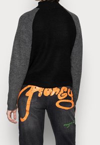 Svart ribbad tröja med grå ärmar, hög halsringning och kontrasterande texturer. Mörka jeans med stor orange "Honey"-tryck och gröna accenter.