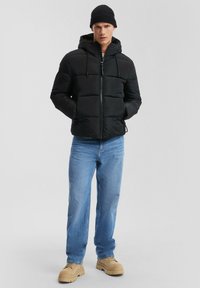 Chaqueta de plumas negra con capucha, que presenta paneles acolchados horizontales, cierre de cremallera y dos bolsillos laterales. Se lleva con jeans azul y zapatos beige.