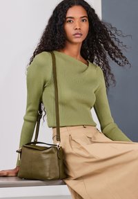 Jeune femme aux longs cheveux bouclés portant un pull vert côtelé et une jupe beige, assise sur un banc avec un sac à main en cuir vert olive.