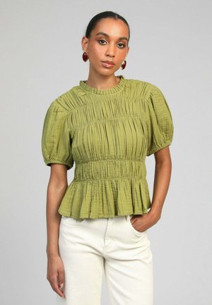 Femme portant un chemisier vert froncé à manches courtes et un pantalon blanc, avec des boucles d’oreilles ovales rouges, debout devant un fond gris uni.