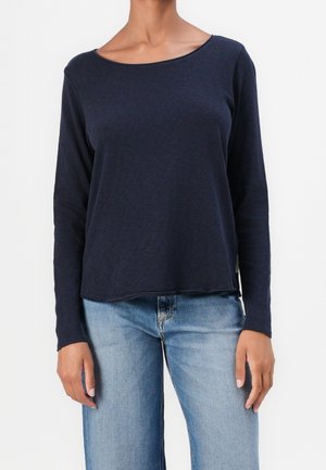 Person trägt ein marineblaues Langarmshirt mit rundem Ausschnitt und hellblaue Jeans, steht mit entspannt an den Seiten hängenden Armen.