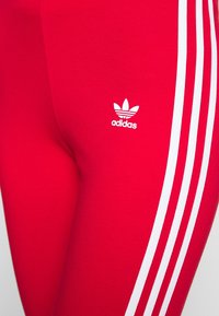 Röda träningstights gjorda av elastiskt material, med tre vita ränder längs sidan och en liten vit Adidas-logotyp på underbenet.
