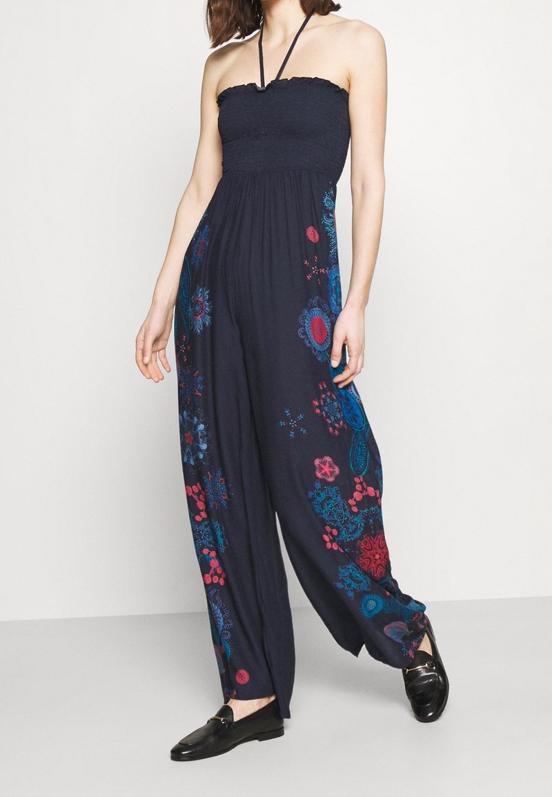 Marinblå storhetslös jumpsuit utan axelband med blomsterdesign i rött och turkos, smockad bodice och vid byxdelsdesign. Texturerat tyg med flödande passform.