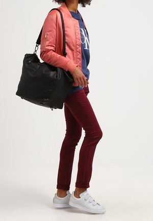 Osoba v růžové bombarďákové bundě, modrém tričku s grafikou, burgundských kalhotách, bílých teniskách a se velkou černou taškou přes rameno.