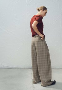 Pull rouge à manches courtes associé à un pantalon large à carreaux beige, avec une taille haute et des poches frontales. Des chaussures foncées complètent la tenue.