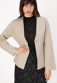 Beige pletený kardigan s hlubokým V-výstřihem a jedním zlatým knoflíkem. Má dlouhé rukávy a vypasovaný střih, kombinovaný s černou vzorovanou sukní.