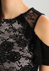 Czarna koronkowa bluzka z misternymi wzorami kwiatowymi, okrągłym dekoltem oraz wycięciami na ramionach, ozdobiona teksturowanym detalem.
