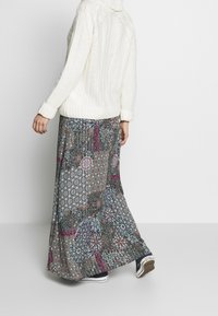 Maxi rok heeft een patronenontwerp in verschillende kleuren, gecombineerd met een crèmekleurige chunky gebreide trui. Schoeisel omvat zwart-witte sneakers.