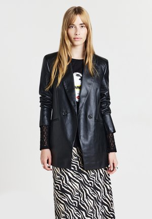 Donna con lunghi capelli biondi che indossa un blazer di pelle nera, maniche in pizzo nero, una maglietta grafica nera e una gonna a stampa zebre su uno sfondo uniforme.